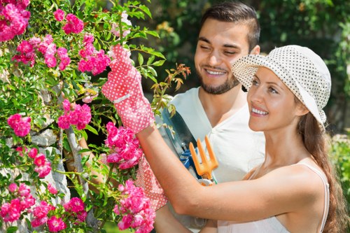 A smiling young couple in a lush front garden during daytime, engaged in gardening activities amidst vibrant pink flowering bushes and green foliage. The woman, wearing a white sunhat and gardening gloves, is tending to the bushes with a small hand rake, while the man, dressed in a light-colored shirt, stands close beside her, observing and supporting her work. The garden features a well-maintained lawn, with dense shrubbery and flowering plants creating a colourful natural setting. Behind them, taller trees provide partial shade, and the scene is illuminated by natural daylight, suggesting a pleasant weather day. This outdoor space exemplifies a tidy, landscaped residential garden ideal for garden maintenance and enhancement, reflecting the expertise of Gardening Services Shoreditch in creating inviting, well-kept outdoor environments in the London postcode area, including areas like Shoreditch.