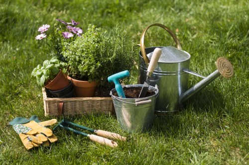 A close-up view of a well-maintained garden area featuring a lush, green lawn with vibrant grass blades. In the foreground, there is a collection of gardening tools and supplies, including a pair of yellow gardening gloves, a small hand trowel, a hand rake, and pruning shears, all resting on the grass. To the left, a woven basket contains several potted plants with varying foliage, some with purple and green leaves, suggesting a mixture of flowering and leafy plants typical of a cultivated garden. Behind the tools and plants, there is a metal watering can with a wooden handle and a long spout, positioned next to a white-handled garden fork lying flat on the grass. The background emphasizes the natural outdoor setting, with the vibrant green lawn extending beyond the gardening supplies, indicating a cared-for, outdoor landscaped space that might be part of a residential garden in Shoreditch. The scene suggests readiness for garden maintenance and planting, aligning with professional gardening services offered in the area.