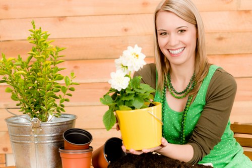 The image shows a smiling woman sitting outdoors in a garden, holding a small yellow flower pot containing a healthy green plant with white flowers. In front of her on a wooden surface, there are empty plant pots of various sizes, including a small black pot and an orange terracotta pot. To her left, a larger potted shrub with bright green leaves is placed in a metallic container. The background features a natural outdoor environment with a wooden fence or wall, indicating a private garden space. The scene is well-lit with natural daylight, suggesting good weather conditions, and the overall setting emphasizes gardening activities associated with garden maintenance, planting, and outdoor beautification visible through the array of pots and healthy plants, consistent with professional gardening services in Shoreditch area.